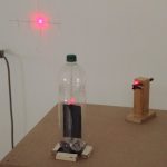 Table top with pop-bottle containing magnet, card and sequin deflecting laser light onto a wall